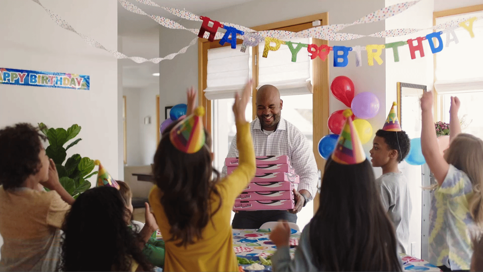 A smiling man carrying pink Hungry Howie’s pizza boxes into a children’s birthday party. The kids in front of the man are jumping and cheering at the arrival of pizza.