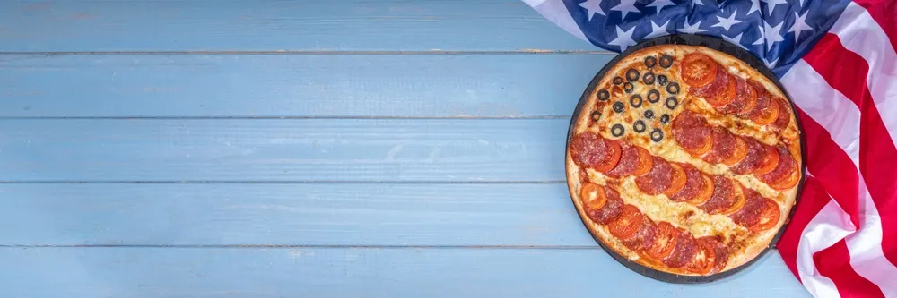 A pizza with pepperoni and black olives with an American flag in the background.